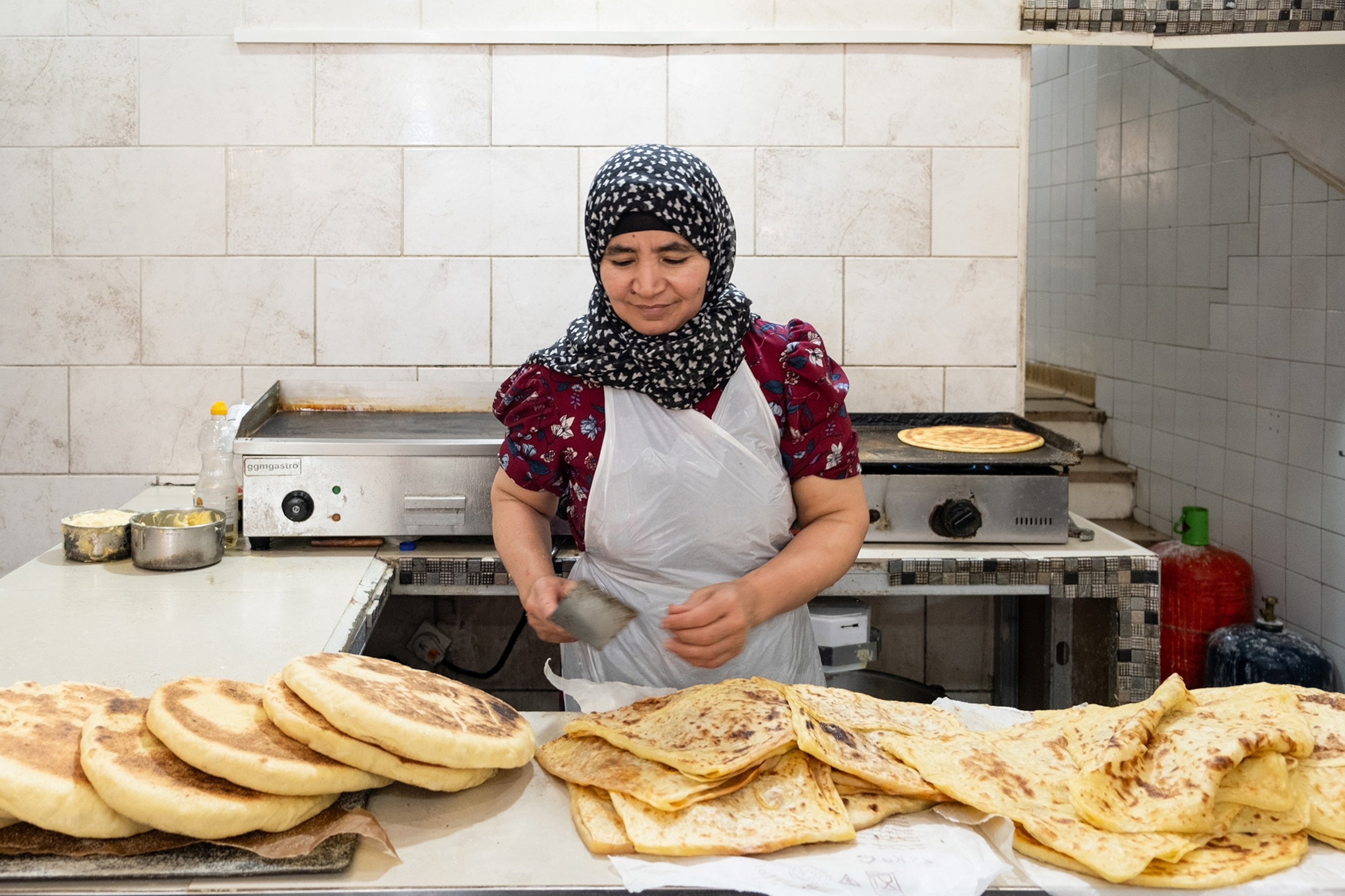 A woman in a hijab hand shaping and baking flat breads in a simple kitchen.