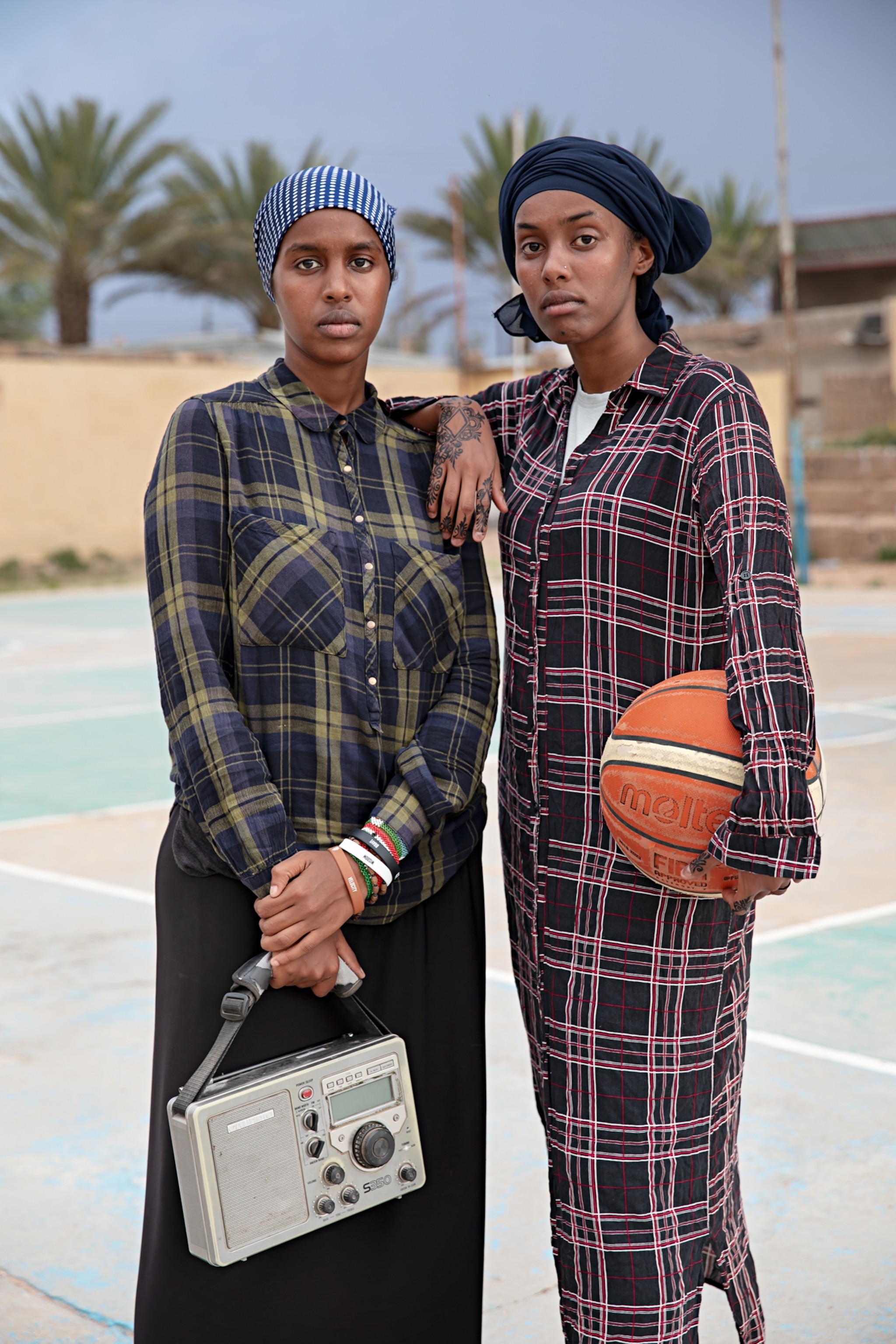 Two women wearing colorful plaid attire stand tall facing forward with a boombox and basketball in their hands.