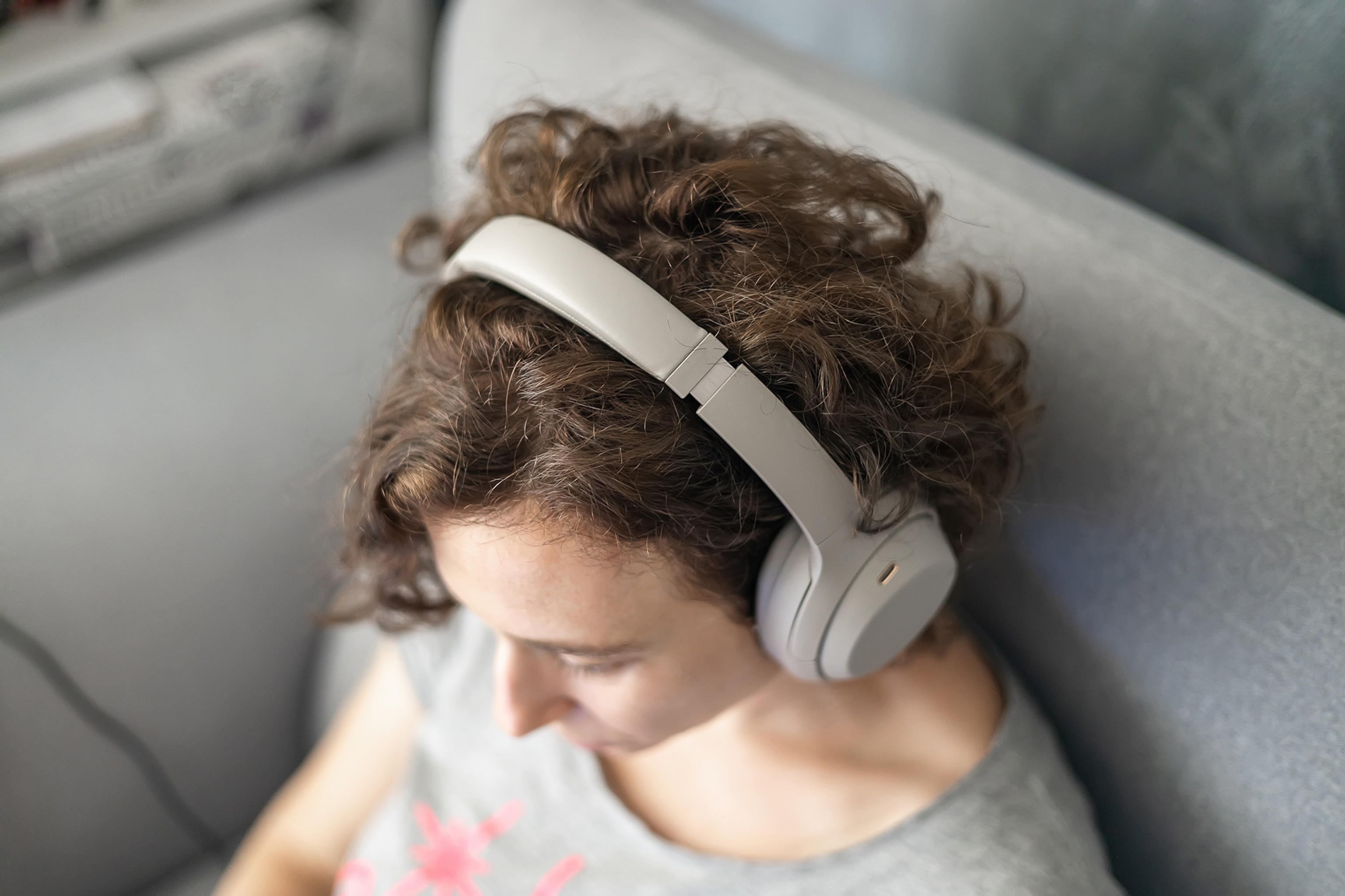 Woman using noise canceling headphones, sitting on gray couch at home.