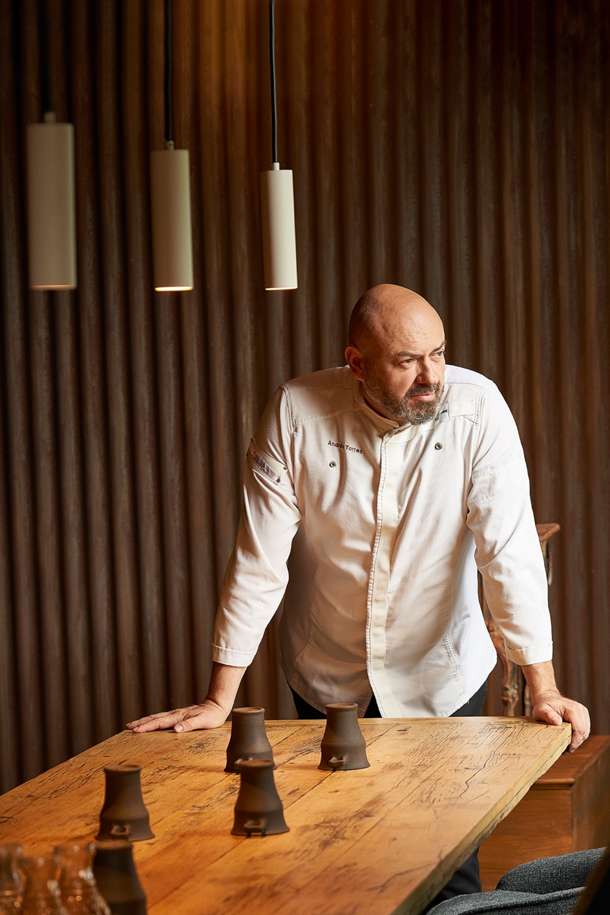 A bald chef in his coat, leaning on the edge of a massive wood table and looking out the wonder with a thoughtful expression.