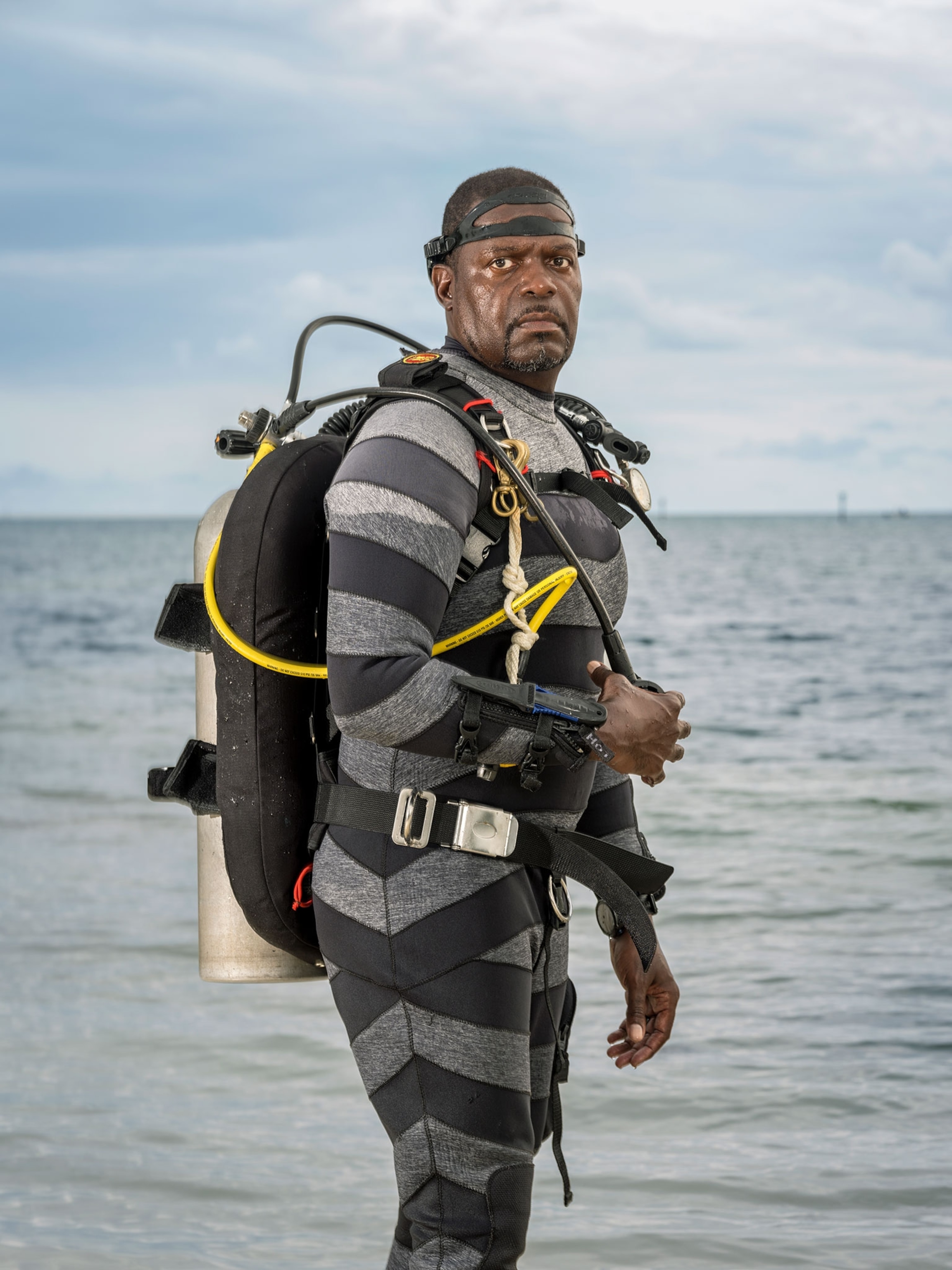 A man wearing scuba gear with an oxygen tank on his back while standing in water up to his waist.