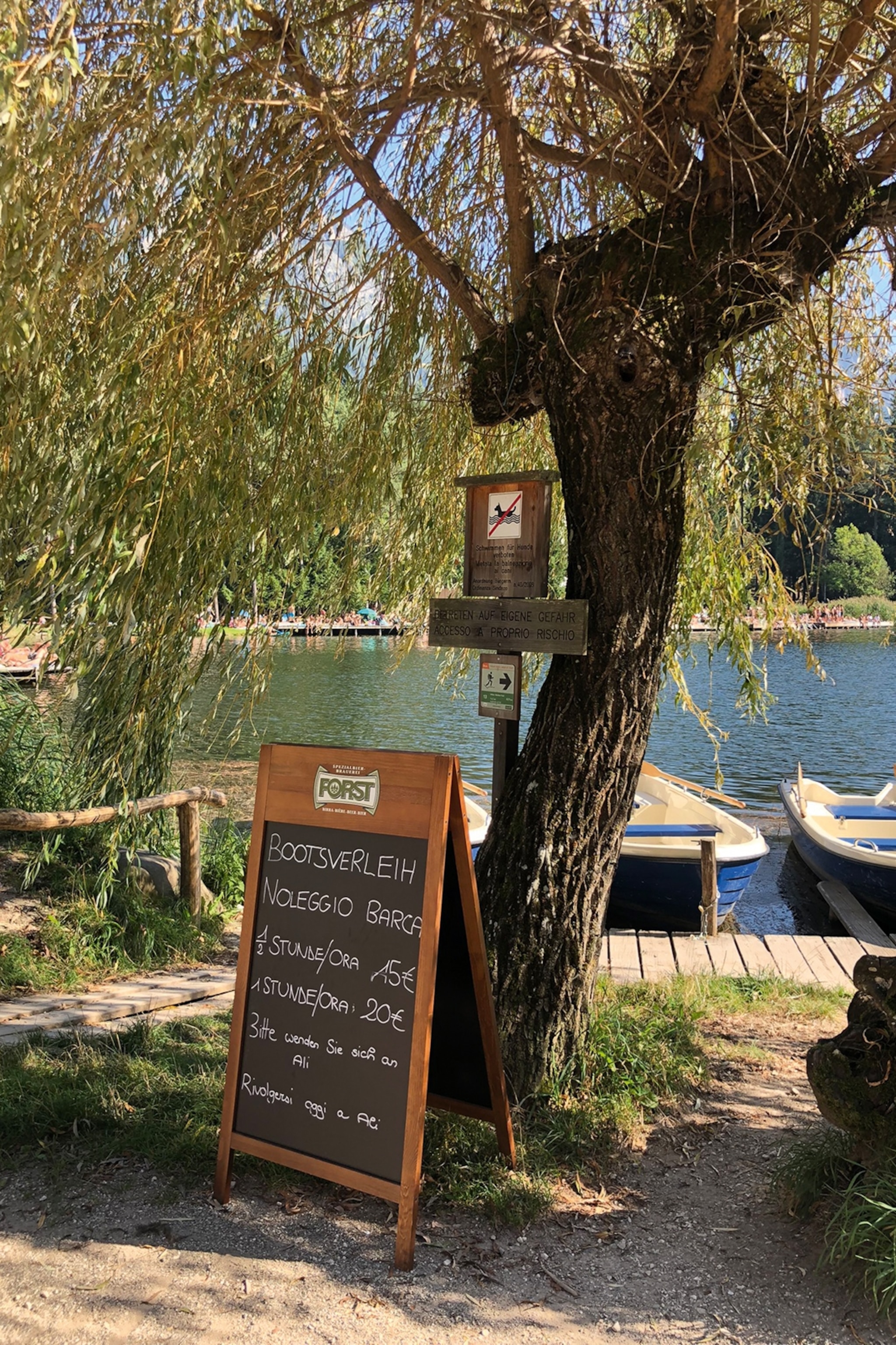 A picture by a swimming lake in the Dolomites. A chalkboard with prices sits beside the water.
