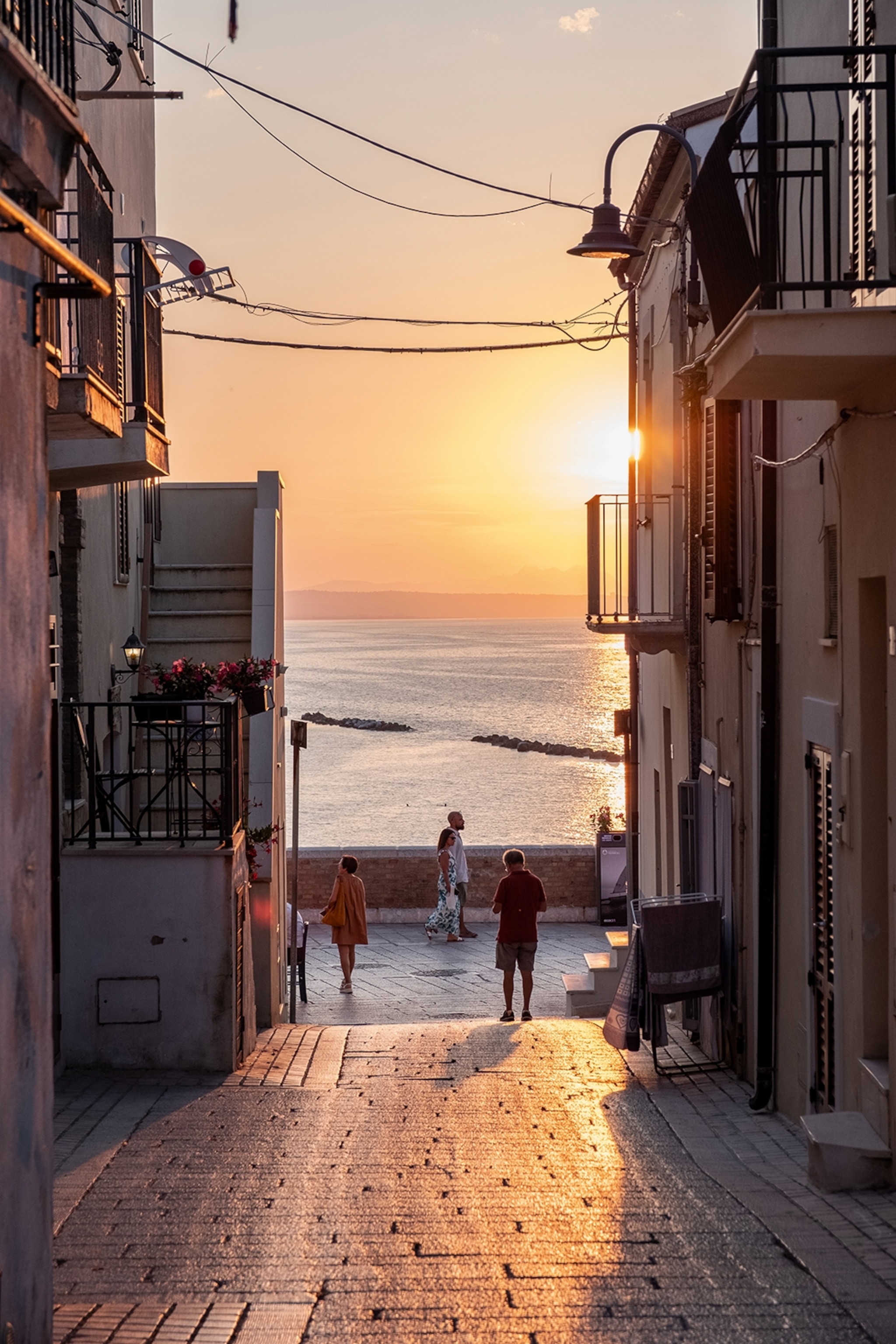 People walk down a narrow street opening up to the sea and a sunset