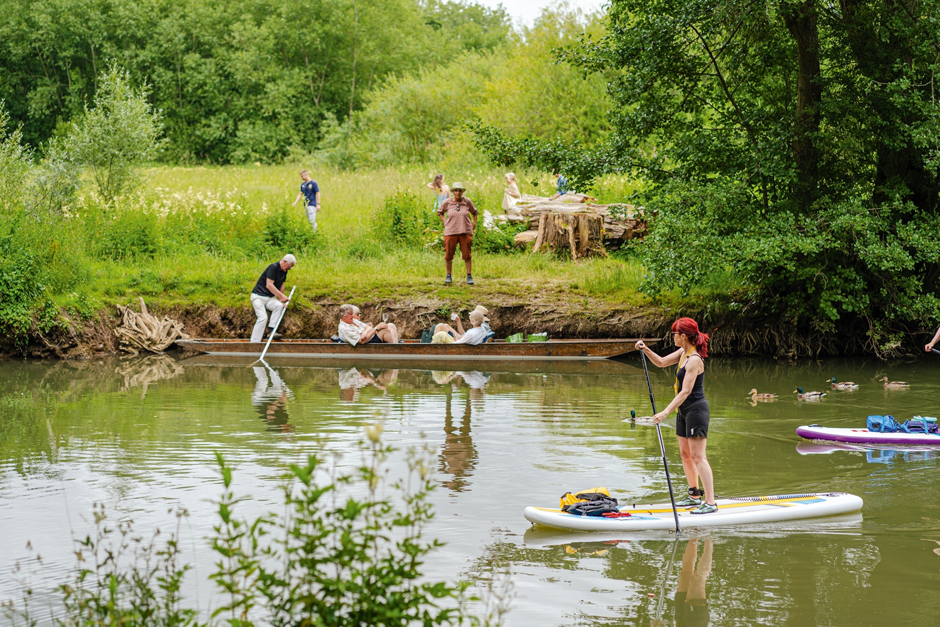 Punter on a boat and person standing on a paddleboard on a river next to a green river bank