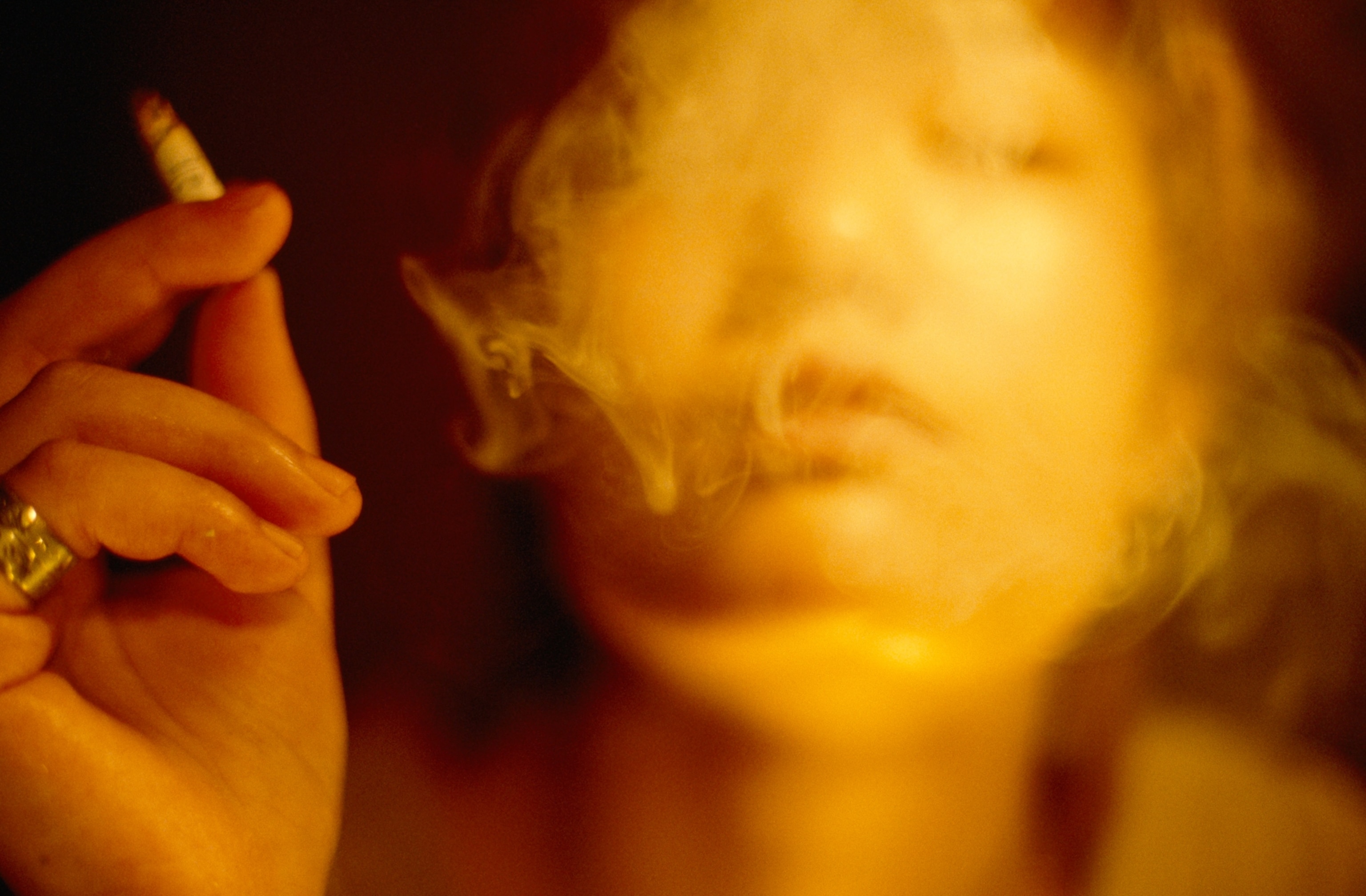 A young woman's face, obscured by smoke as she exhales, a lit cigarette casually held in her hand.