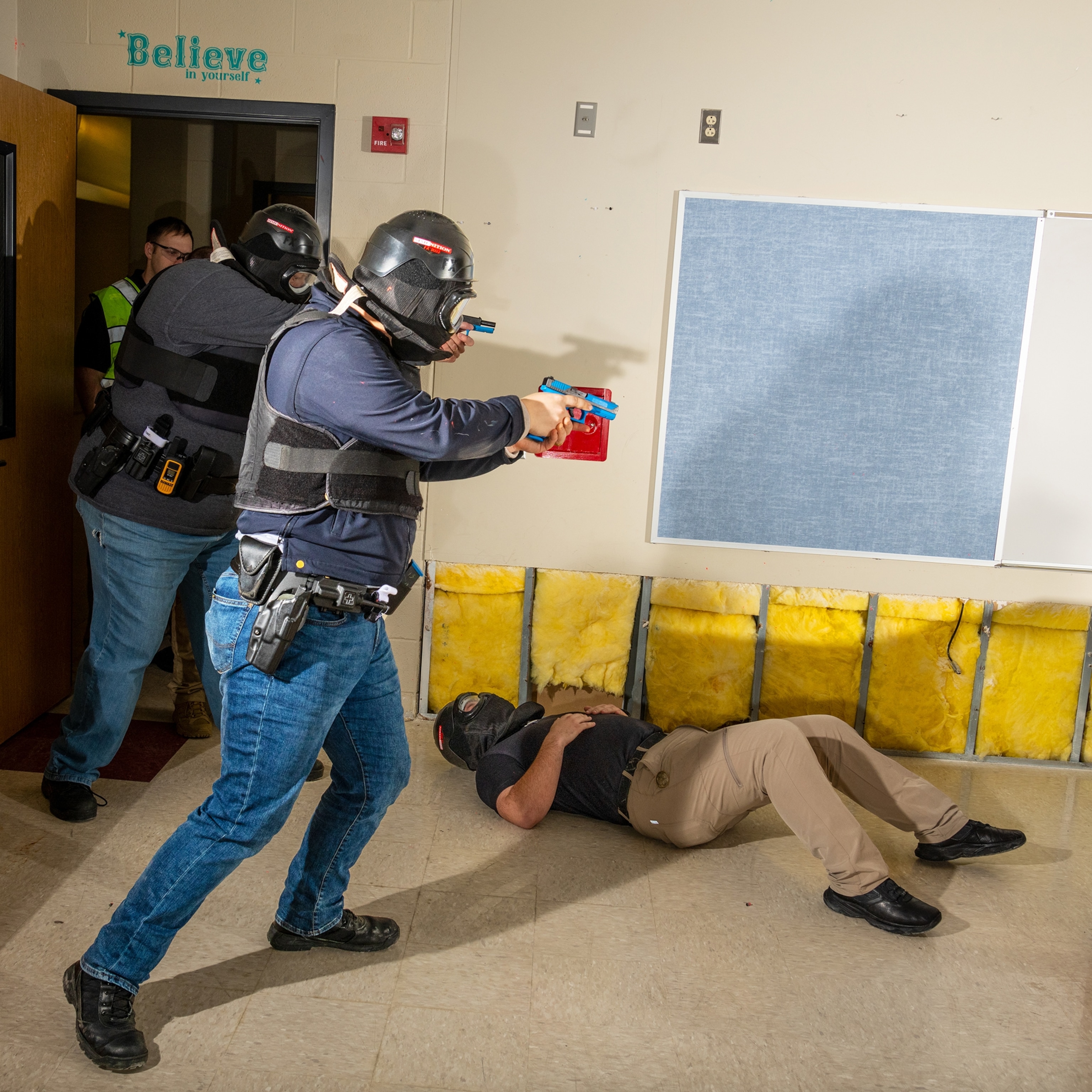 Two men in a classroom wearing safety helmets and protective gear hold out their guns as a another man lays on the ground facing the ceiling.