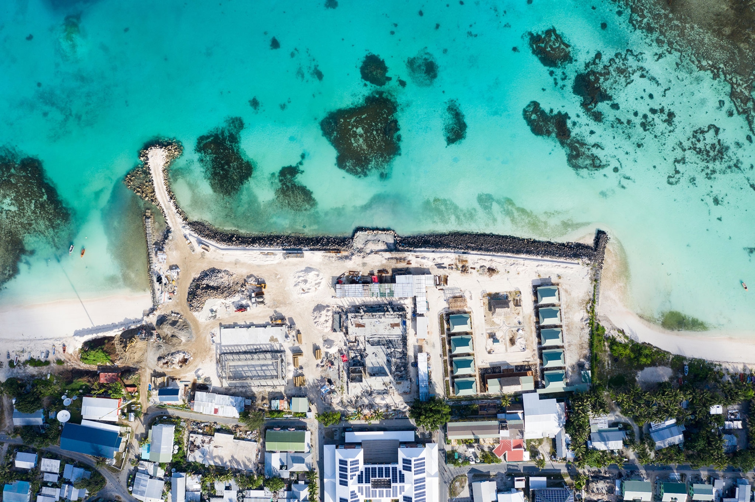An aerial shot from straight above a construction site on Funafuti island shows construction vehicles moving sand to extend the islands land into the ocean. The photo is shot from far above making the many house on the bottom of the frame feel small, while the ocean at the top of the frame fills half of the photo with blue/green water.