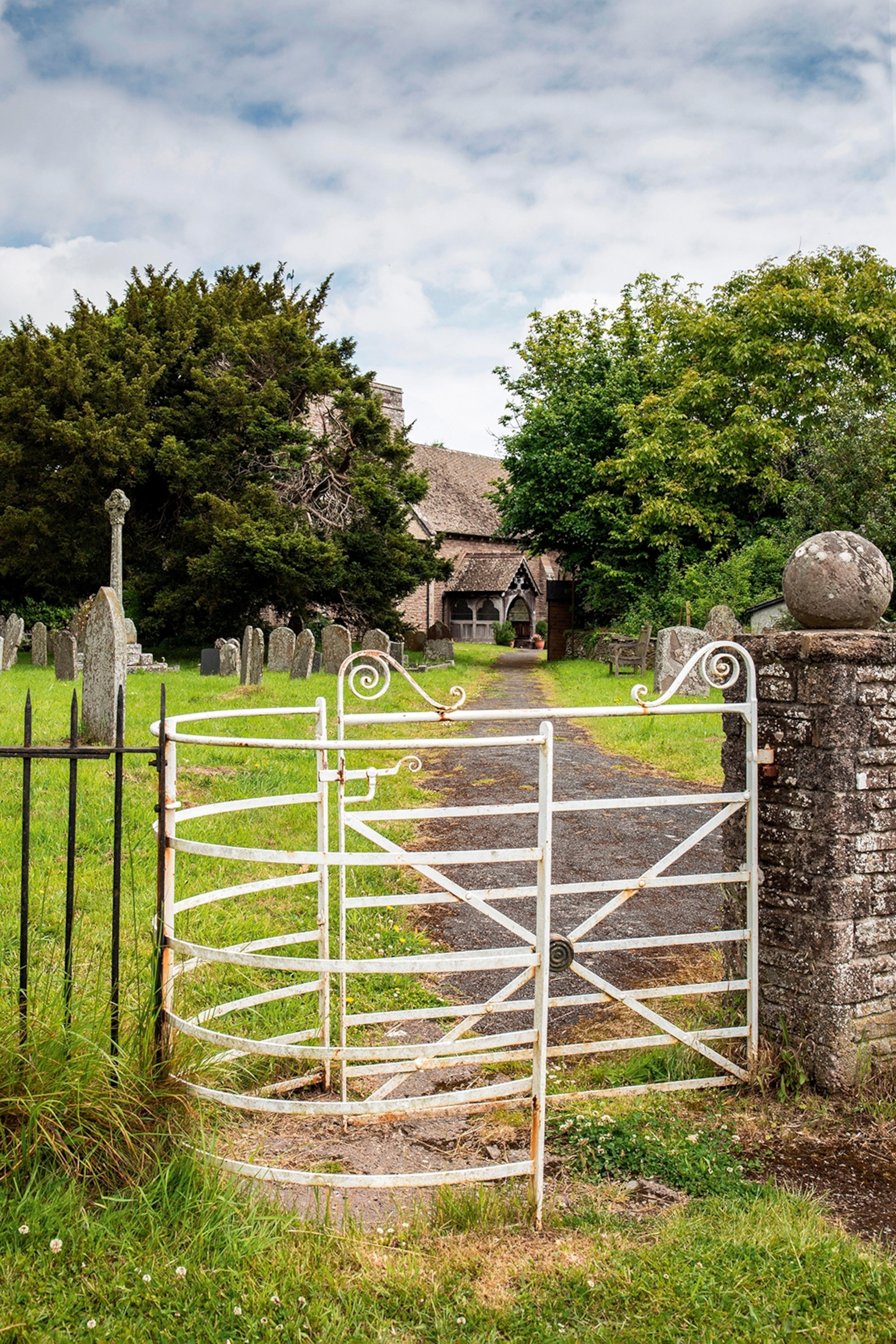 A curved iron gate with a path leading to a medieval church, half hidden by trees.