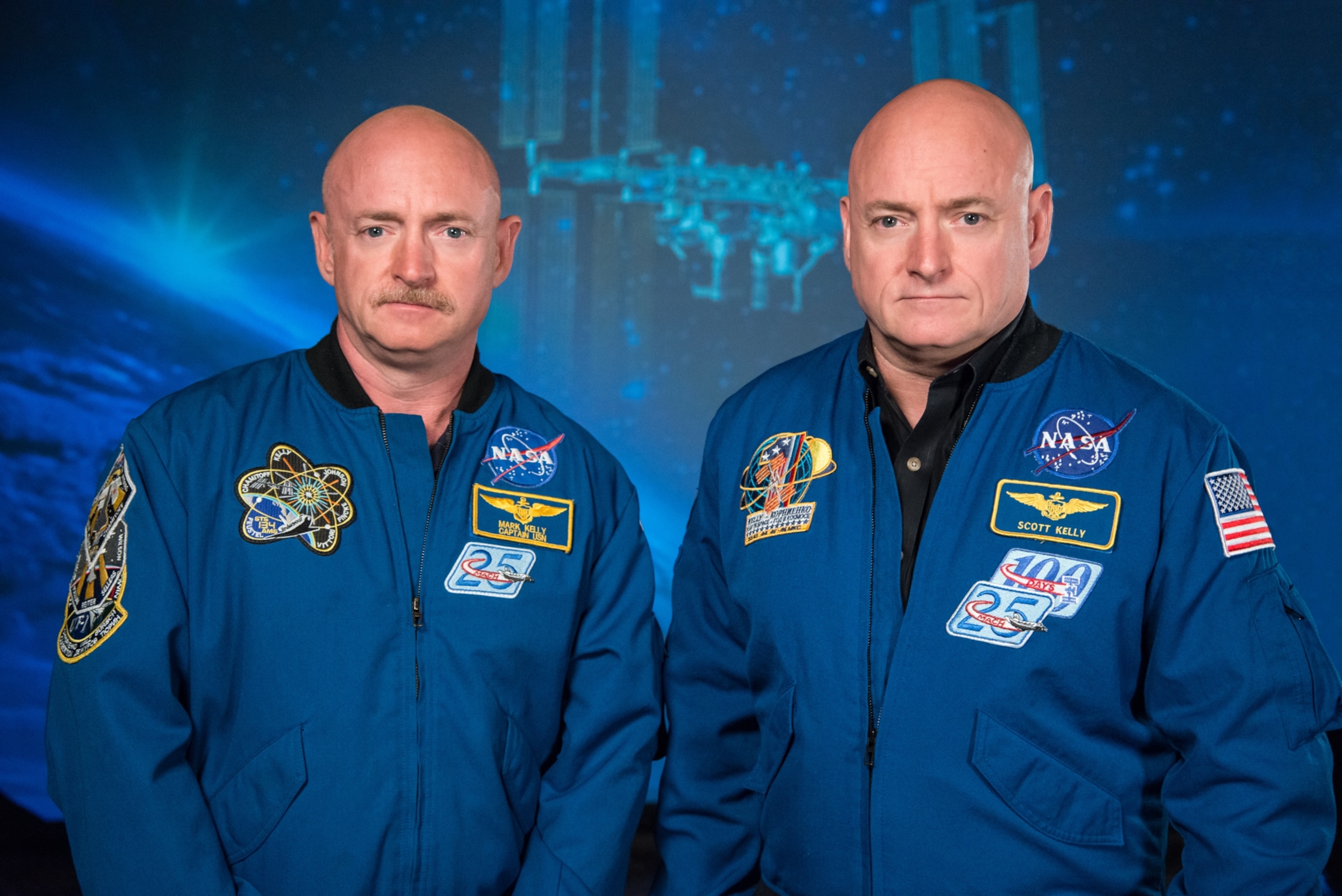 Identical twin brothers, retired Astronaut Mark Kelly and Astronaut Scott Kelly standing side by side in blue jump suits.