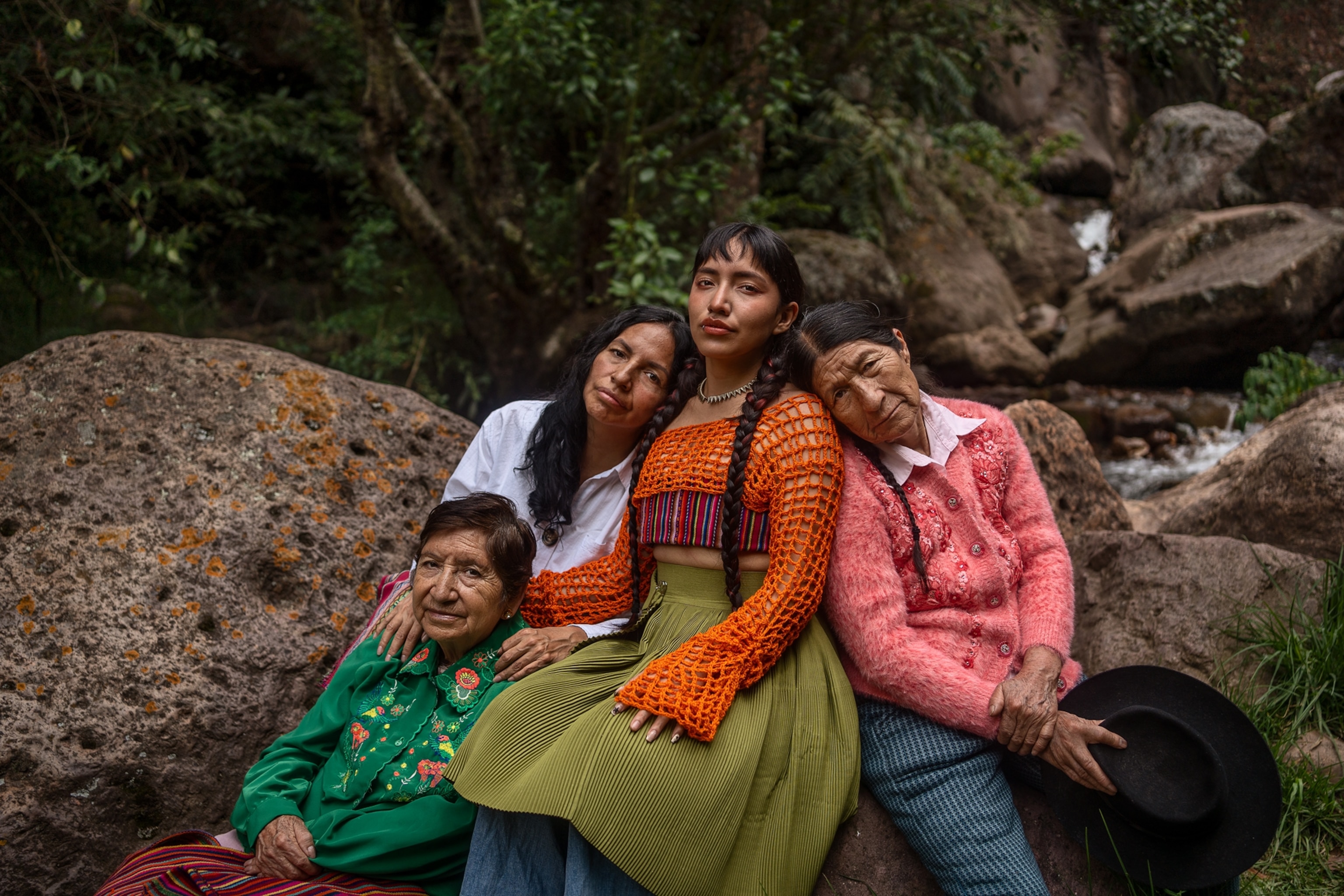 Woman, Flores, is seen sitting on a rock with her mother and two grandmothers sitting on either side of her. They are all seen leaning their heads on her shoulders.