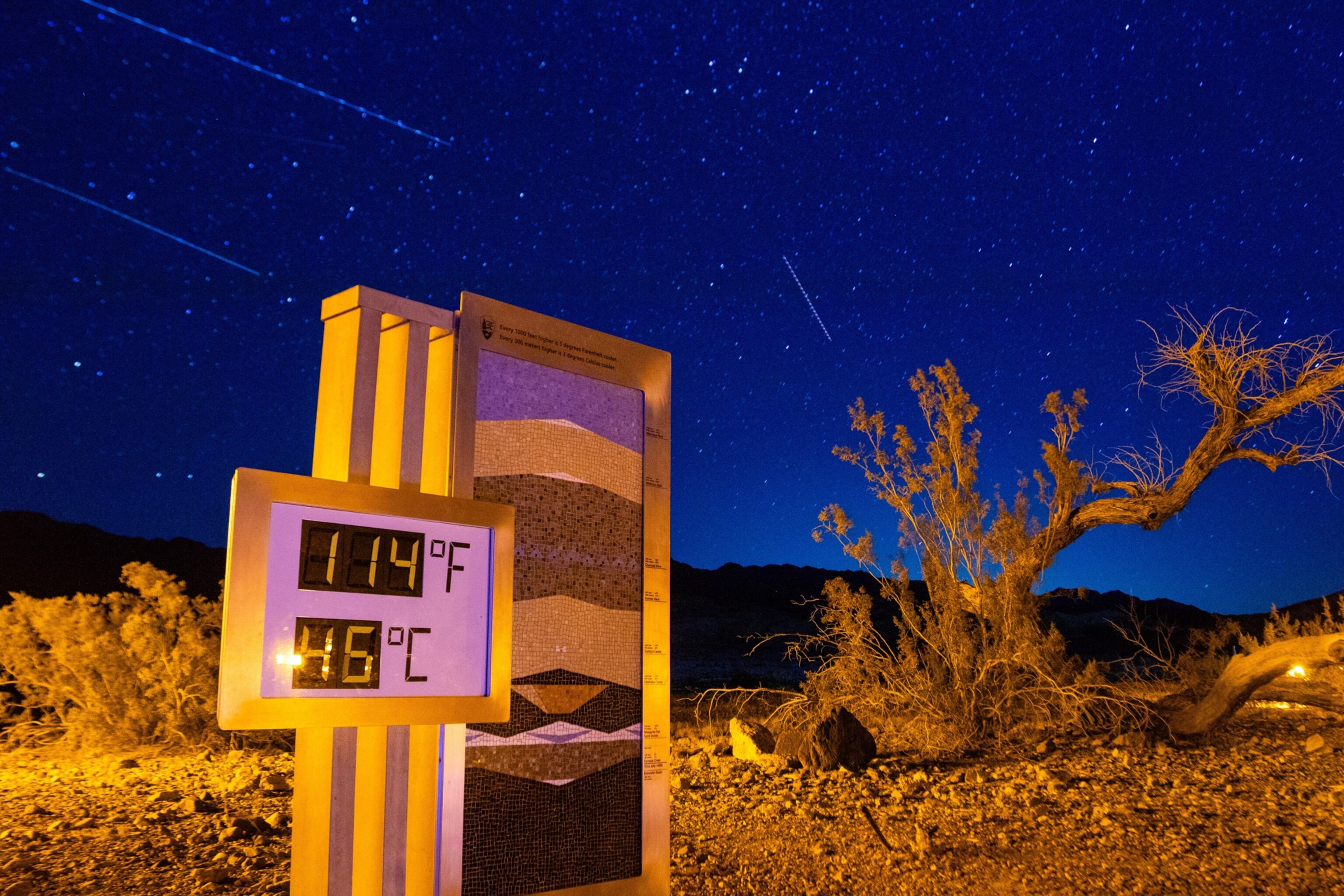A night time view of Death Valley where a sign displaying the temperature reads," 114 degrees Fahrenheit. 45 degrees Celsius" with a starry sky above