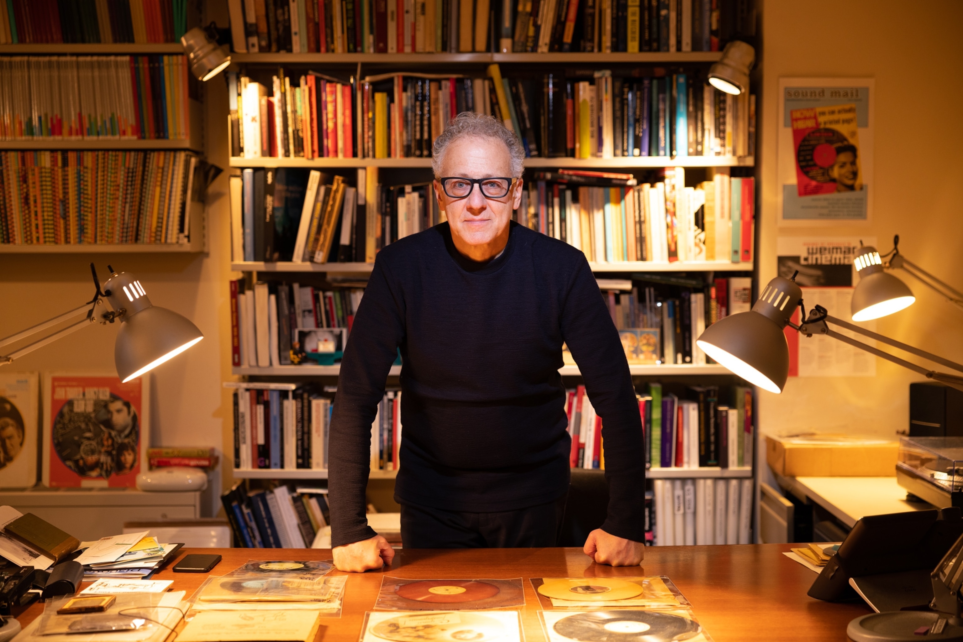 Photo of man with records and books on table and bookshelf behind him.