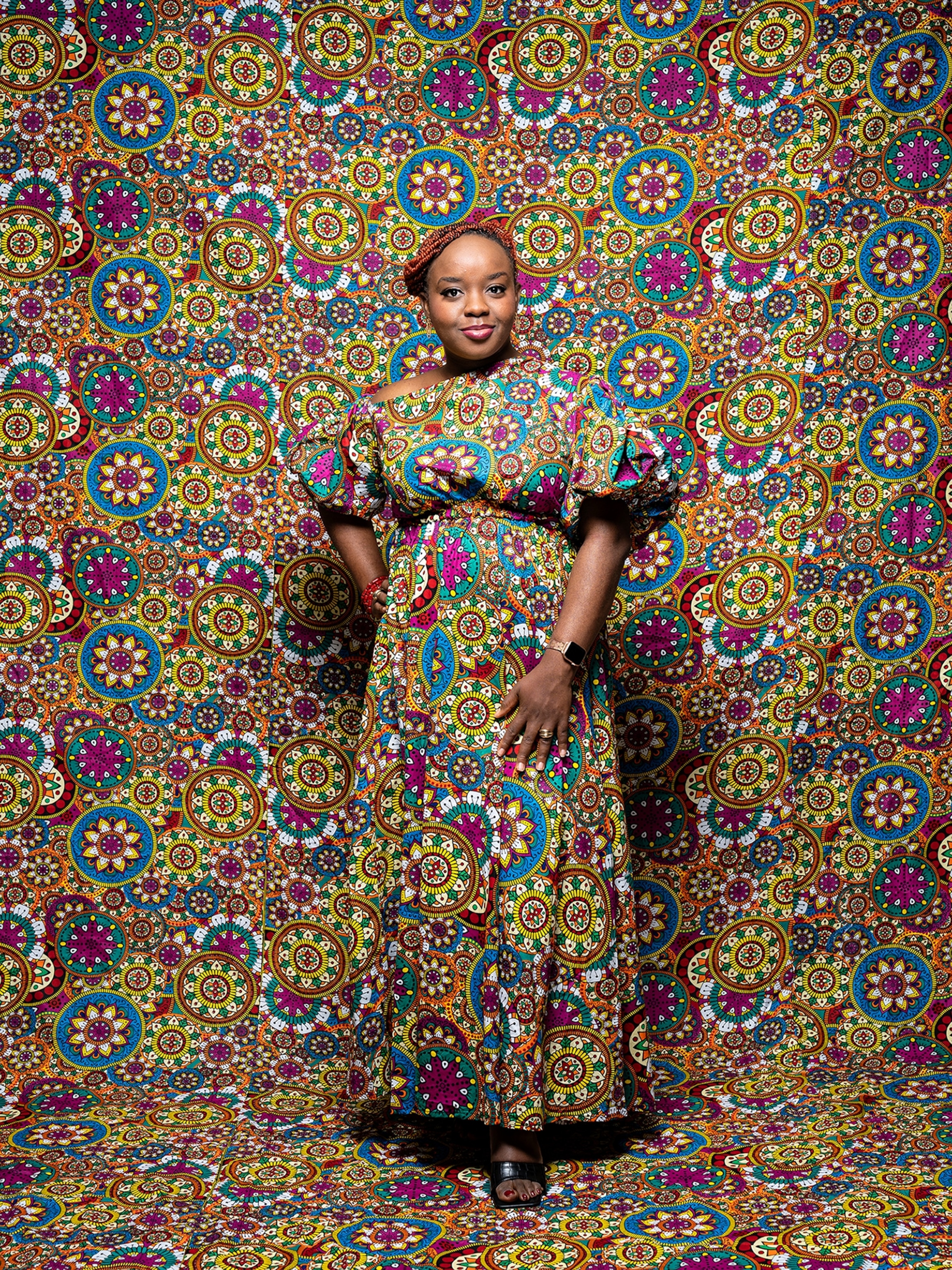 A woman poses for a portrait in a bright and busy patterned dress. Both the floor and wall are covered in the same pattern.