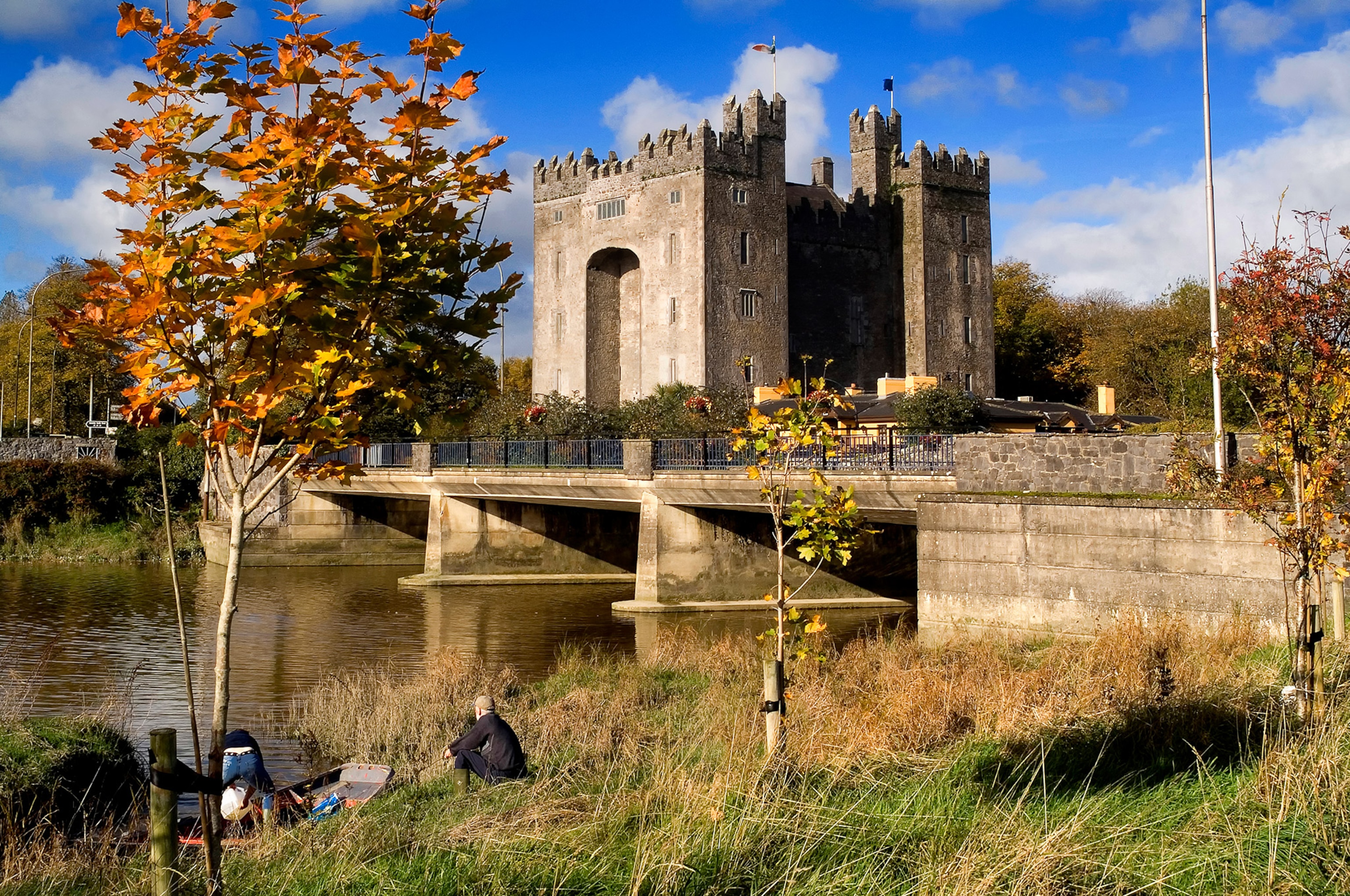 People sit together near the river bank with the castle overlooking the landscape in the background.