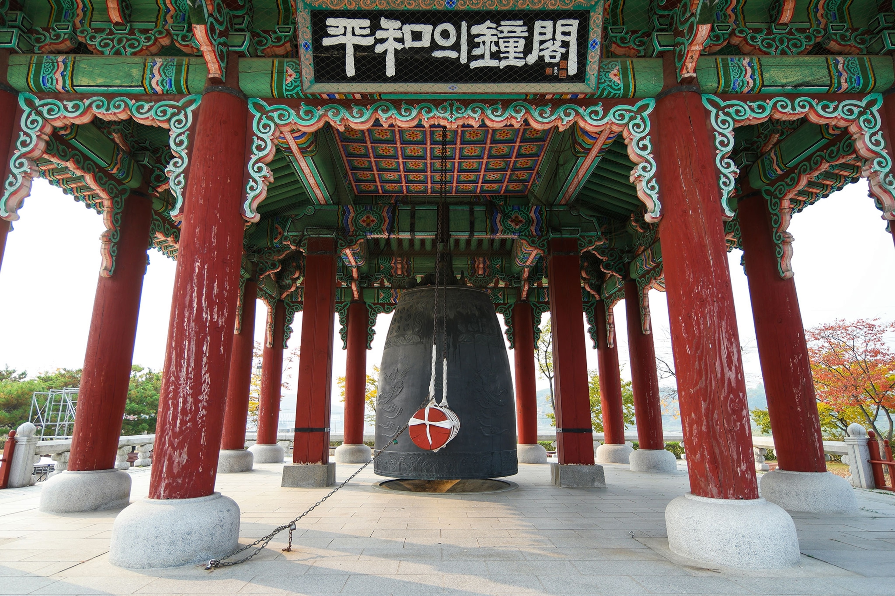 A bell stands under a red and green ornately-decorated pavilion.